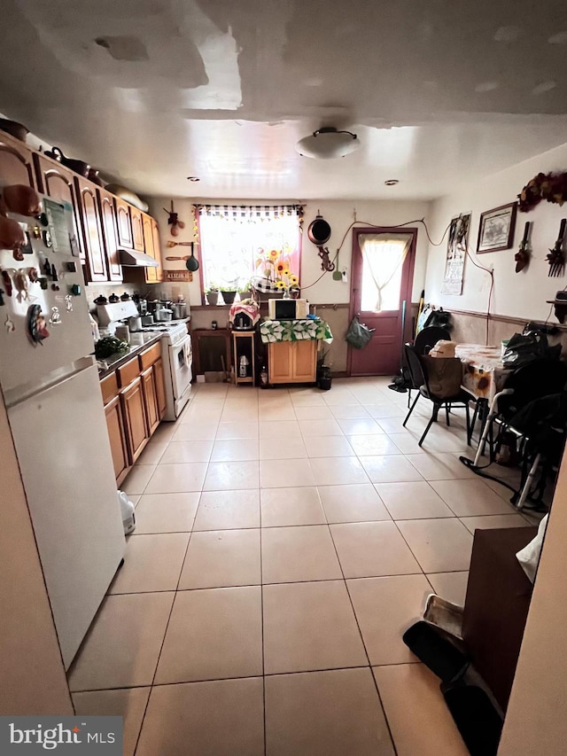 kitchen with light tile patterned floors, under cabinet range hood, white appliances, wainscoting, and brown cabinets