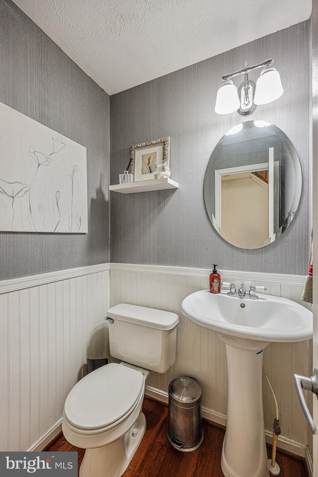 half bathroom featuring a textured ceiling, toilet, a wainscoted wall, wood finished floors, and wallpapered walls