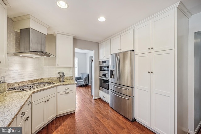 kitchen with backsplash, appliances with stainless steel finishes, white cabinetry, wall chimney exhaust hood, and hardwood / wood-style flooring