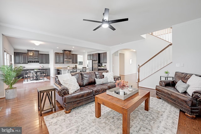 living room featuring crown molding, ceiling fan, stairs, light wood-style floors, and arched walkways