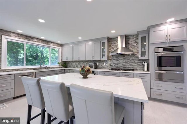 kitchen with a warming drawer, a kitchen bar, gray cabinets, stainless steel appliances, and wall chimney range hood