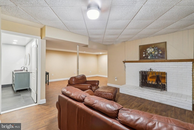 living area featuring a brick fireplace, baseboards, a drop ceiling, and wood finished floors