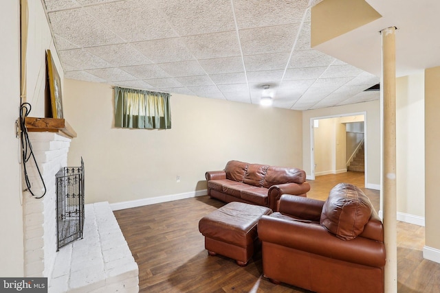 living area with a brick fireplace, a drop ceiling, baseboards, and wood finished floors