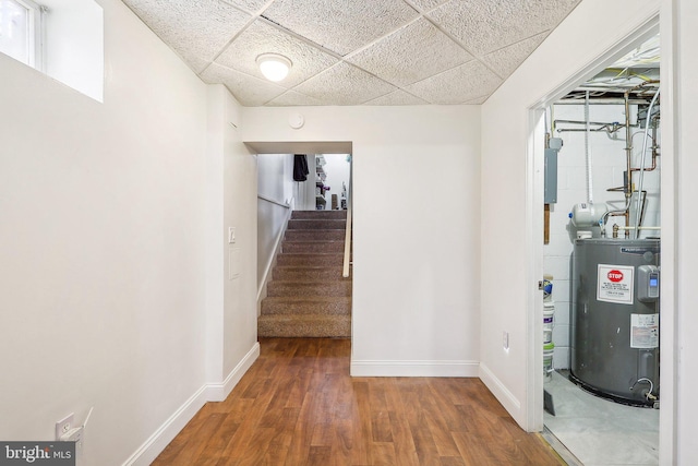 hall featuring a paneled ceiling, wood finished floors, baseboards, water heater, and stairs