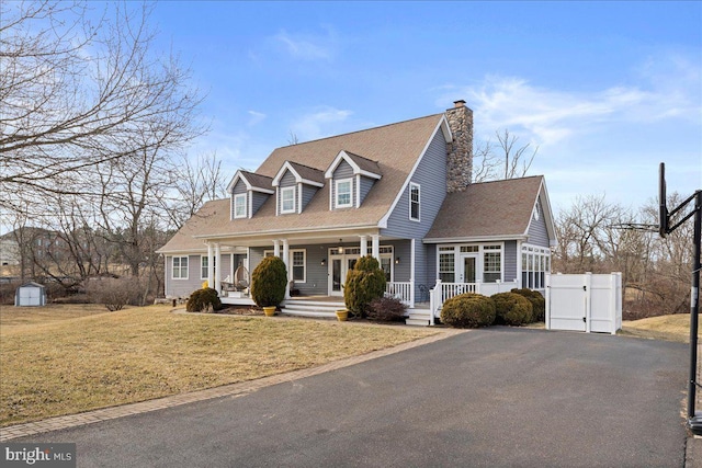 cape cod house featuring covered porch, driveway, a gate, a front lawn, and a chimney