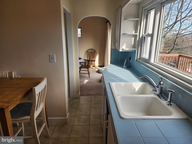 kitchen with tile counters, arched walkways, tile patterned floors, open shelves, and a sink