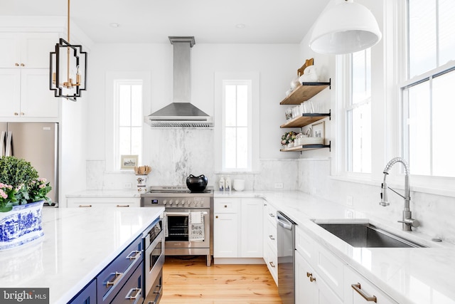 kitchen with blue cabinets, stainless steel appliances, a sink, white cabinets, and wall chimney range hood