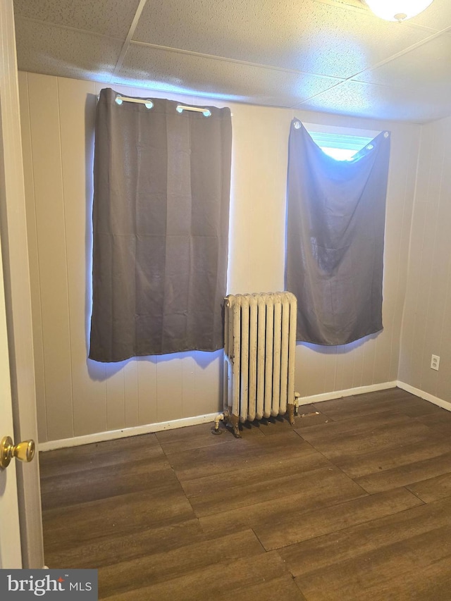 empty room with a drop ceiling, radiator heating unit, baseboards, and wood finished floors