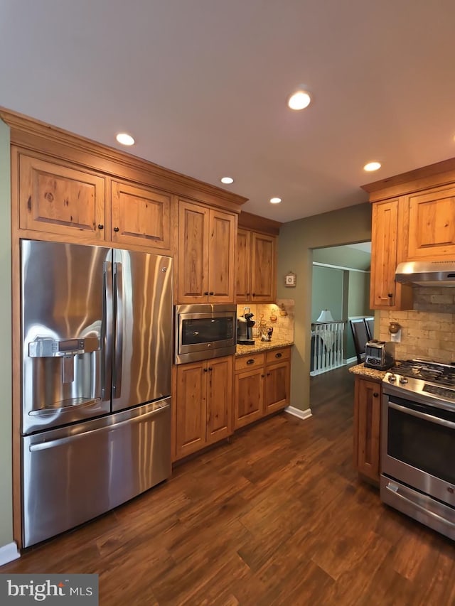 kitchen featuring tasteful backsplash, appliances with stainless steel finishes, dark wood-type flooring, light stone countertops, and under cabinet range hood