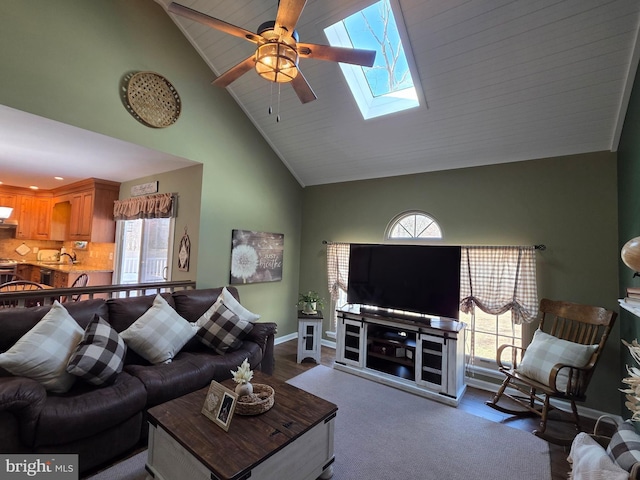 living room featuring ceiling fan, high vaulted ceiling, a skylight, and baseboards