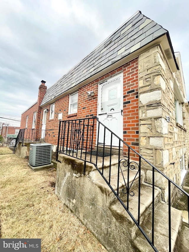 exterior space featuring a shingled roof, central AC unit, mansard roof, and brick siding