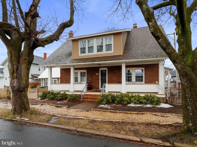 view of front facade with a shingled roof, a chimney, fence, a porch, and brick siding