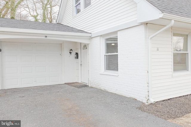 view of exterior entry featuring an attached garage, driveway, a shingled roof, and brick siding