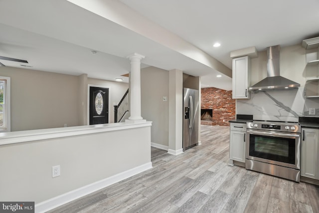 kitchen featuring light wood-style floors, appliances with stainless steel finishes, wall chimney exhaust hood, dark countertops, and decorative columns