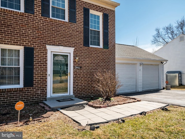 view of exterior entry featuring brick siding, an attached garage, roof with shingles, and driveway