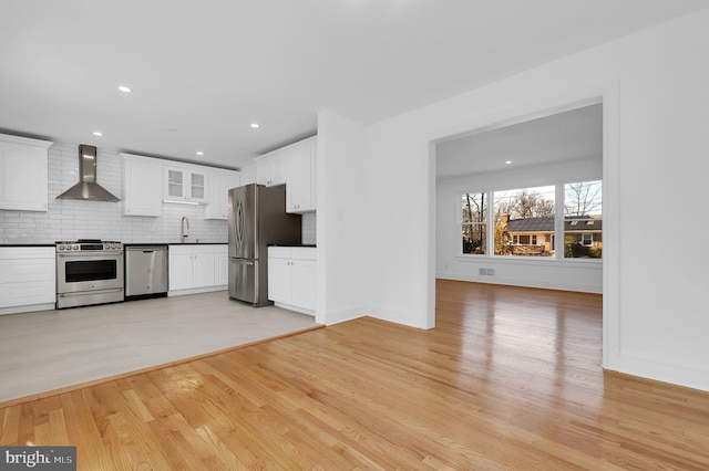 kitchen with light wood finished floors, decorative backsplash, white cabinets, appliances with stainless steel finishes, and wall chimney range hood