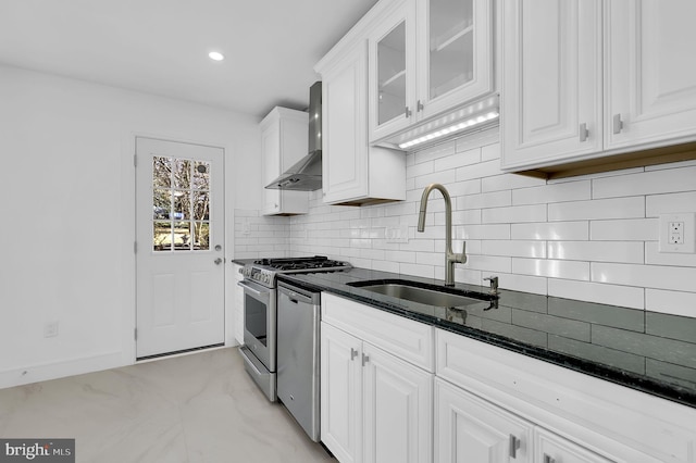 kitchen with dark stone counters, a sink, appliances with stainless steel finishes, white cabinetry, and wall chimney range hood