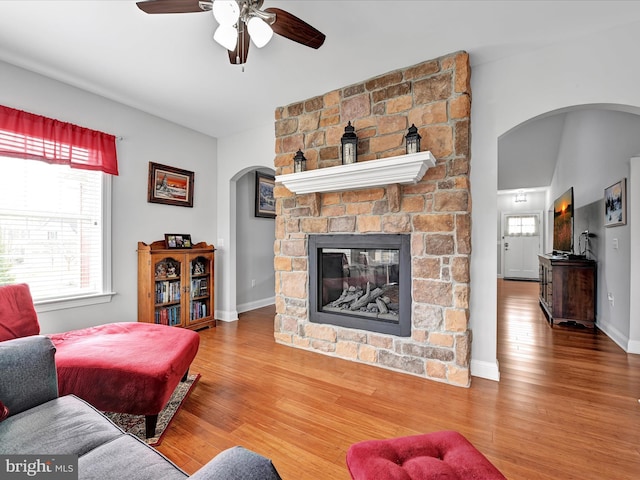 living area featuring arched walkways, ceiling fan, a fireplace, wood finished floors, and baseboards