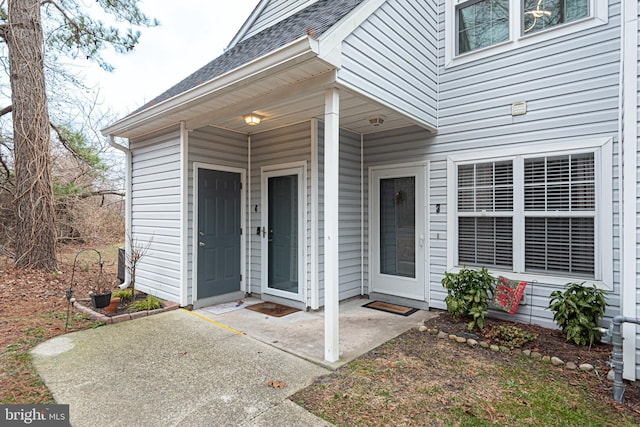 view of exterior entry with a shingled roof and a patio