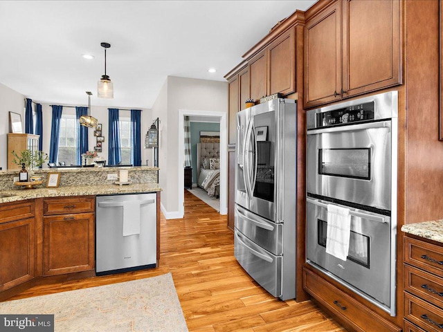 kitchen featuring appliances with stainless steel finishes, brown cabinets, light stone counters, hanging light fixtures, and light wood-style floors