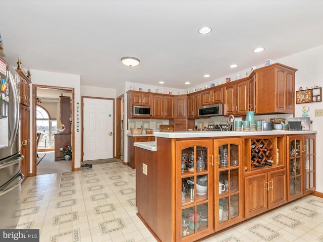 kitchen with a peninsula, light countertops, appliances with stainless steel finishes, brown cabinetry, and glass insert cabinets