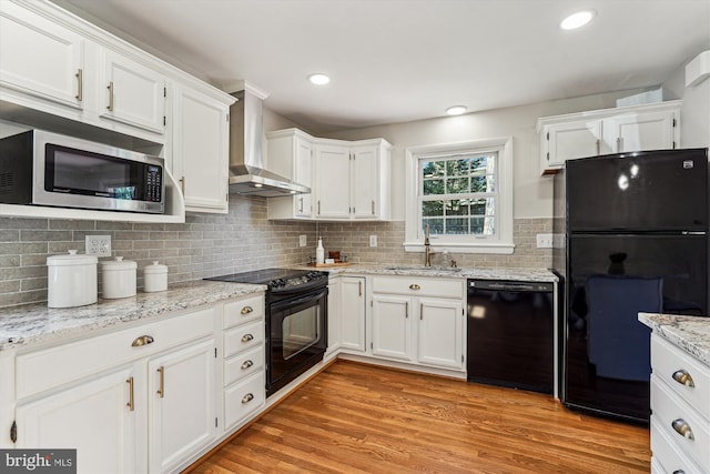 kitchen featuring a sink, black appliances, white cabinetry, wall chimney range hood, and light wood-type flooring