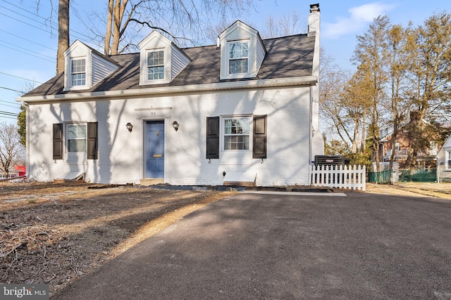 cape cod-style house featuring roof with shingles, a chimney, fence, and brick siding