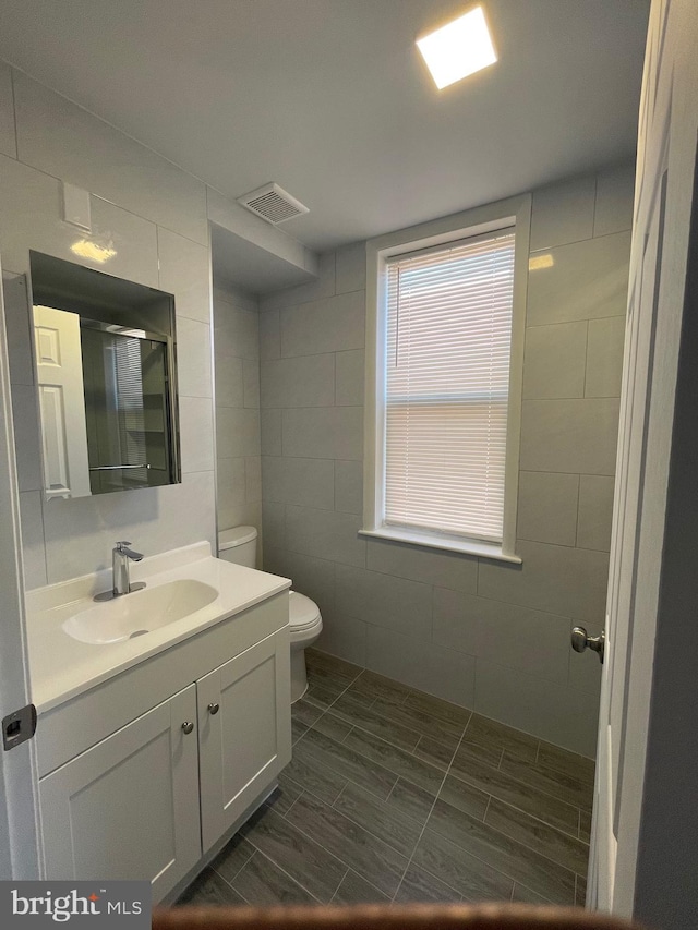 bathroom with a stall shower, visible vents, toilet, vanity, and tile walls