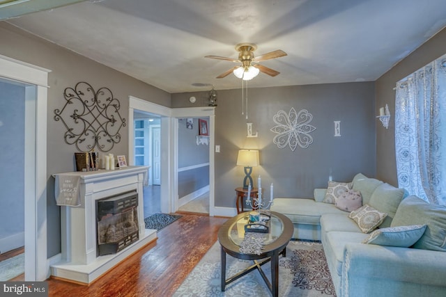 living area with a glass covered fireplace, ceiling fan, baseboards, and wood finished floors