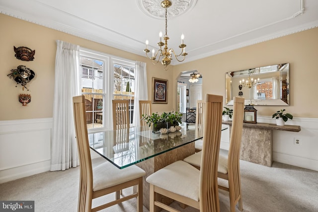 carpeted dining area featuring ceiling fan with notable chandelier, ornamental molding, and wainscoting