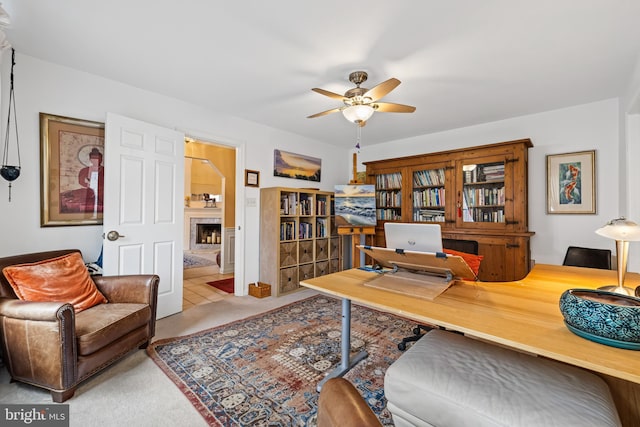 home office with carpet floors, a fireplace with raised hearth, and a ceiling fan