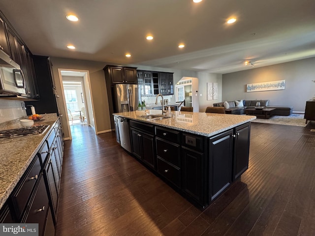 kitchen with a kitchen island with sink, dark wood-type flooring, a sink, open floor plan, and appliances with stainless steel finishes