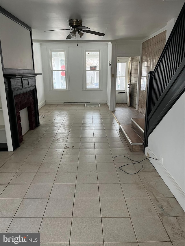 unfurnished living room featuring light tile patterned floors, a ceiling fan, baseboards, stairway, and a brick fireplace