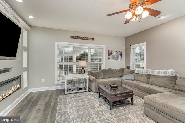 living area with a glass covered fireplace, wood finished floors, baseboards, recessed lighting, and a ceiling fan
