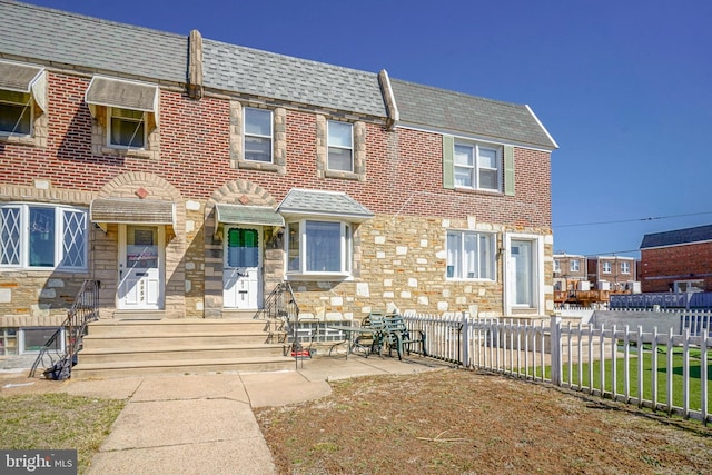 townhome / multi-family property with stone siding, brick siding, roof with shingles, and fence