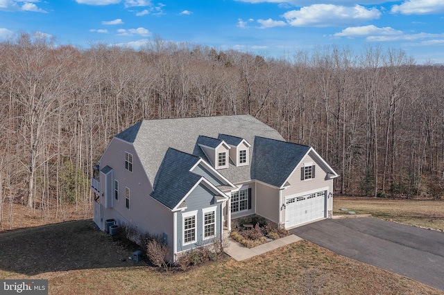 view of front of property with a shingled roof, a view of trees, a garage, cooling unit, and driveway