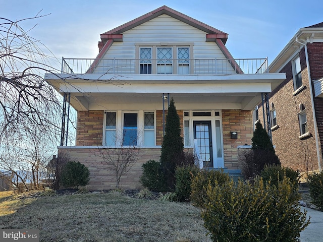 view of front of property with stone siding, covered porch, a balcony, and a gambrel roof