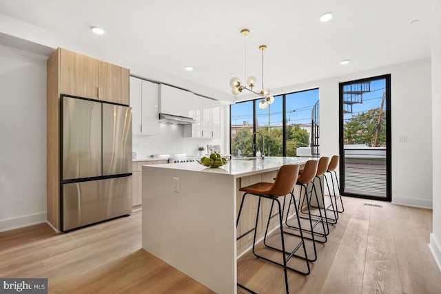 kitchen featuring under cabinet range hood, light wood-style floors, light countertops, freestanding refrigerator, and an island with sink