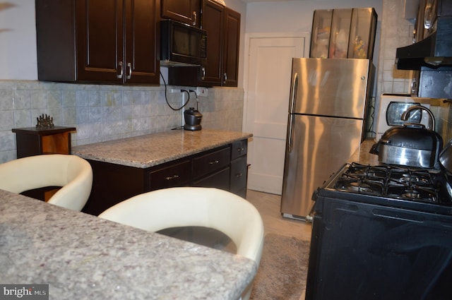 kitchen featuring light stone counters, under cabinet range hood, dark brown cabinets, black appliances, and tasteful backsplash