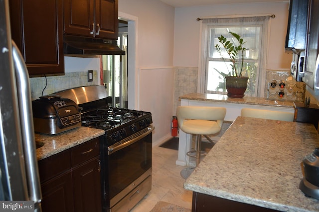 kitchen with light stone counters, appliances with stainless steel finishes, wainscoting, dark brown cabinets, and under cabinet range hood