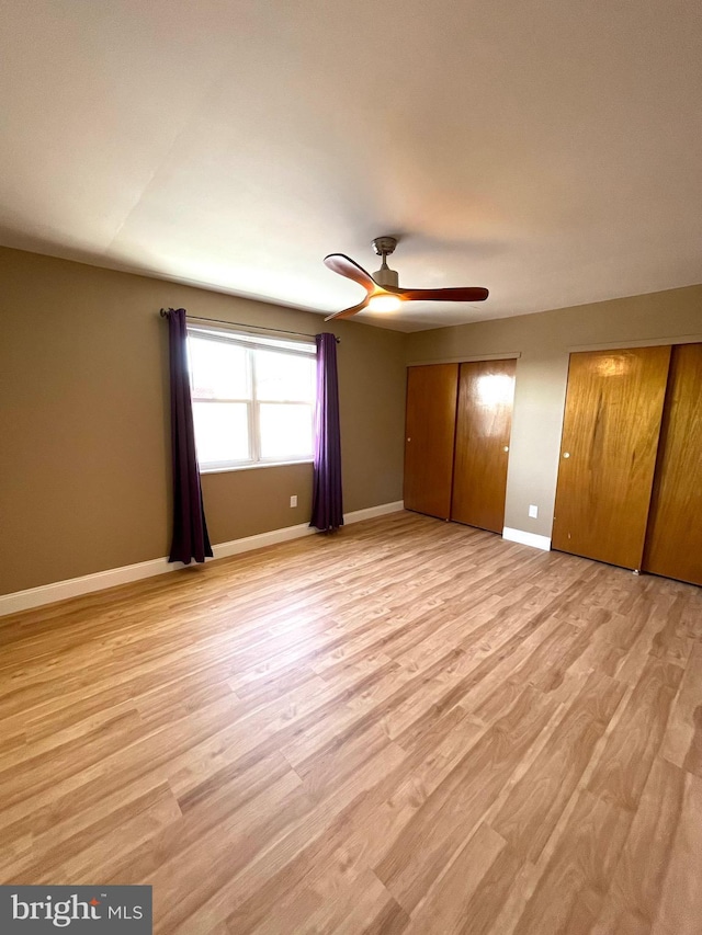 unfurnished bedroom with light wood-type flooring, a ceiling fan, baseboards, and two closets