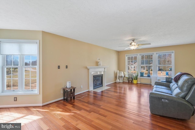 living area featuring a textured ceiling, a fireplace with flush hearth, wood finished floors, a ceiling fan, and baseboards