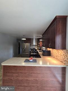 kitchen with a peninsula, wall chimney exhaust hood, stainless steel fridge with ice dispenser, light countertops, and dark brown cabinets