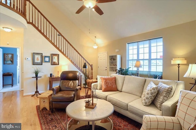 living room with baseboards, ceiling fan, stairs, light wood-style floors, and high vaulted ceiling