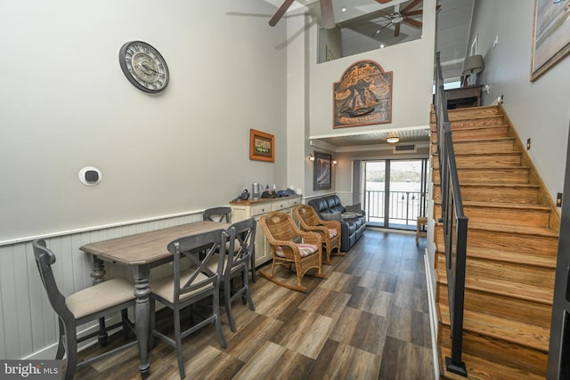 dining area featuring a ceiling fan, a towering ceiling, a wainscoted wall, wood finished floors, and stairs