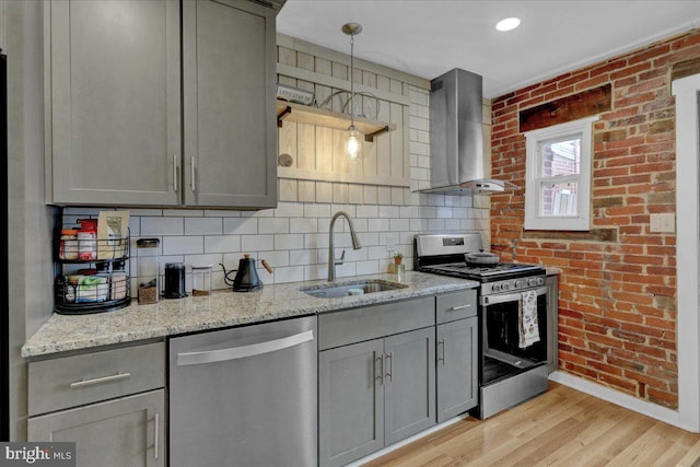 kitchen with gray cabinets, appliances with stainless steel finishes, a sink, wall chimney range hood, and brick wall