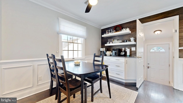 dining area featuring ceiling fan, a decorative wall, a wainscoted wall, dark wood-type flooring, and crown molding