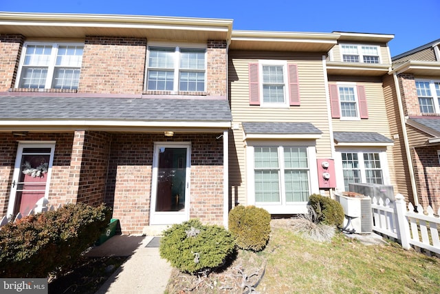 multi unit property featuring roof with shingles, fence, central AC unit, and brick siding