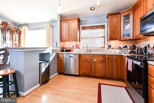 kitchen with light wood-style flooring, a sink, appliances with stainless steel finishes, brown cabinetry, and glass insert cabinets