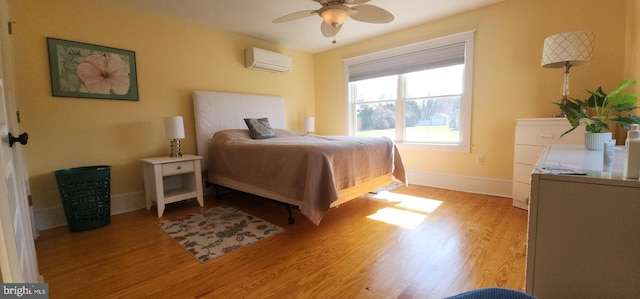 bedroom with a ceiling fan, a wall unit AC, light wood-style floors, and baseboards